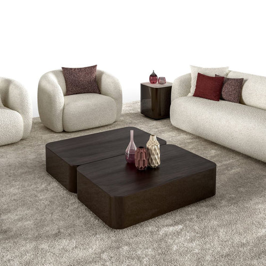 A modern living room setup featuring a dark brown wooden coffee table with two stacked square sections, styled with decorative vases. The room includes a cream boucle sofa with red and purple accent pillows, a matching cream boucle armchair, and a small dark side table on a textured gray rug.