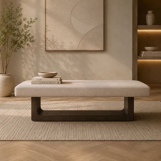A modern upholstered bench with a light beige fabric seat and a dark wood base, styled in a warm, minimalist living room setting.