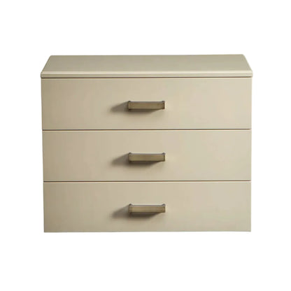 A three-drawer nightstand or storage cabinet in a warm cream-beige finish with slim metallic drawer pulls, shown against a plain white background.